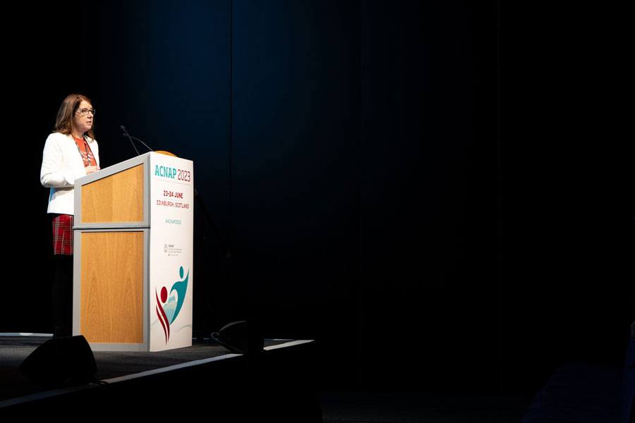 Speaker at ACNAP 2023 stands at a podium in a darkened room