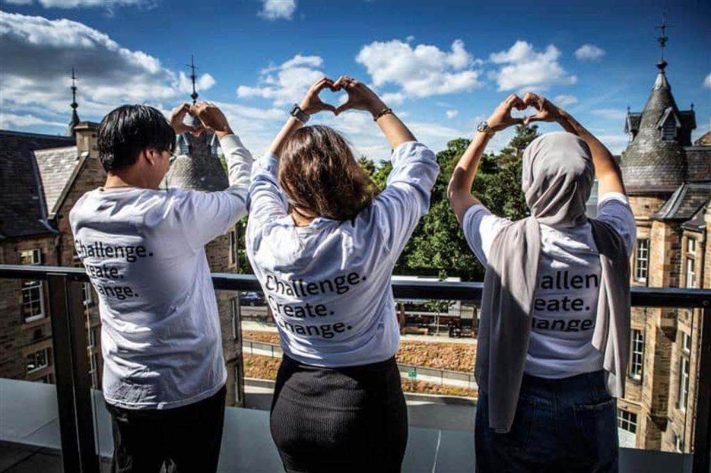 Three people wearing t-shirts with Challenge Create Change on the back make the heart symbol with their hands as they look out over edinburgh