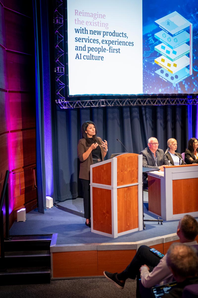 A woman is presenting at a lectern next to a panel. Behind her a slide with the title Reimagine the existing with new products, services, experiences and people-first AI culture